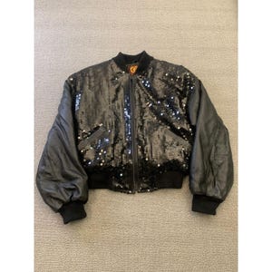 May include: Black sequin bomber jacket with black leather sleeves and a black ribbed collar and cuffs. The jacket has a front zipper closure and two front pockets. The jacket is a cropped style.