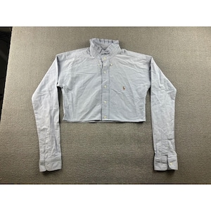 May include: A light blue cropped button-down shirt with long sleeves. The shirt features a ruffled collar and a small embroidered logo on the chest. The sleeves have buttoned cuffs.