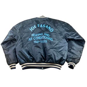 May include: A navy blue satin jacket with white and black striped cuffs and collar. The back of the jacket features the text "JOE FASANO HEATING AND AIR CONDITIONING 752-3555" in light blue lettering.