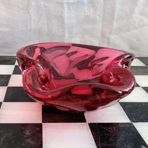 Könnte beinhalten: Eine tiefrote Glasschale mit einem durchscheinenden, organischen und gewellten Rand. Die Schale hat eine glatte, polierte Oberfläche mit verschiedenen Schattierungen von Karmesinrot und Rubin, die eine fließende, asymmetrische Form bilden.