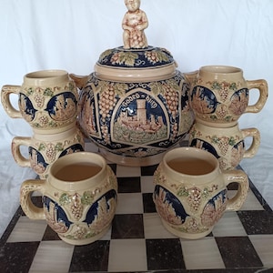 Puede incluir: Un juego de jarras de cerveza de cerámica vintage con una jarra grande con tapa y una figura de querubín en la parte superior, y seis tazas a juego. El juego es de color beige con detalles en azul y marrón, y un diseño de castillo. La jarra dice "Godes-burg".