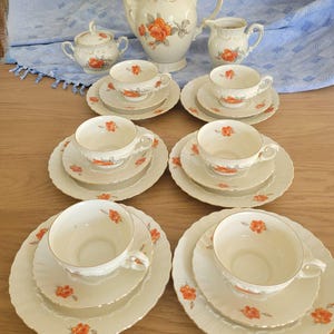 May include: A cream-colored porcelain tea set featuring a delicate orange rose pattern with gold trim. The set includes a teapot with a lid, a sugar bowl with a lid, a creamer, and six teacups with matching saucers and dessert plates.