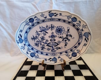 Op de afbeelding: Ovale serveerschaal met een witte basis en een blauw bloemmotief. De schaal heeft een geschulpte rand en wordt ondersteund door een goudkleurige metalen standaard. Het blauwe ontwerp toont bloemen en bladeren.