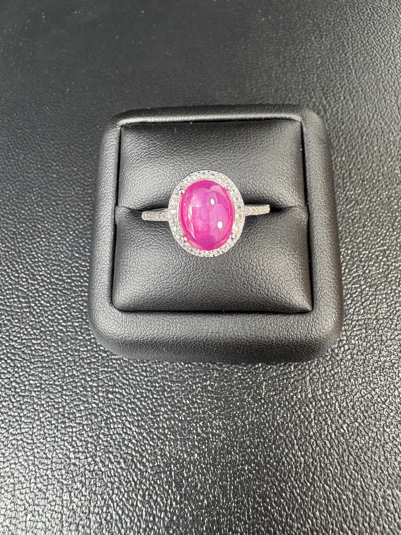 May include: A silver ring featuring a large, oval-cut, vibrant pink gemstone surrounded by a halo of smaller, clear stones. The ring is set on a black display box, showcasing the jewelry's brilliance.