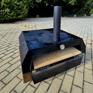 May include: Black outdoor pizza oven with a rectangular shape, a chimney, and a door. The oven has a thermometer and sits on a wooden pallet. The background features green trees and a brick patio.