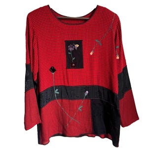 May include: Red long-sleeve top with a black grid pattern and floral embroidery. The top features black panels and a square patch with a flower and butterfly design. The sleeves are a solid red color.
