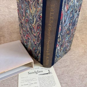 May include: A hardcover book titled Tales of Mystery & Imagination by E.A. Poe with a marbled cover featuring swirls of blue, red, and gold. The spine is brown with gold lettering. A cream-colored slipcase and a Sandglass pamphlet are included.