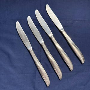 Puede incluir: Cuatro cuchillos de mantequilla plateados con mangos curvos y diseños de estrellas. Las hojas son lisas. Los cuchillos están dispuestos en diagonal sobre un fondo azul oscuro. Un juego de cubiertos.