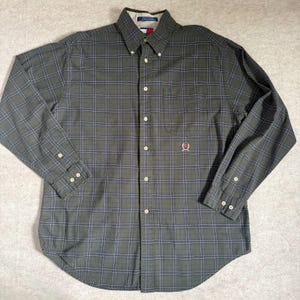 May include: A long-sleeve button-down shirt in olive green with a dark blue and white grid pattern. The shirt features a button-down collar, a single chest pocket with a small embroidered logo, and light-coloured buttons along the front placket.