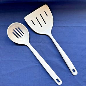 May include: Two white kitchen utensils: a slotted spoon and a spatula. The spoon has a rounded head with four slots, and the spatula has a rectangular head with three slots. Both have long handles with a hole at the end for hanging.