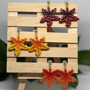 Puede incluir: Tres pares de pendientes con forma de hoja en colores otoñales. Los pendientes se exhiben en un pequeño caballete de madera. El par superior es de color burdeos intenso, el par del medio es de color amarillo a naranja y el par inferior es de color naranja a rojo.
