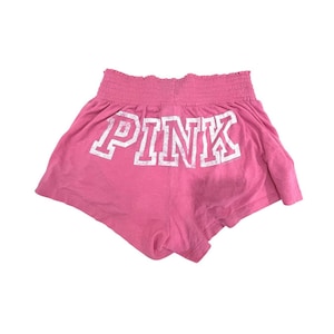 May include: Pink athletic shorts in a vibrant pink hue. The shorts feature a shirred waistband and the word "PINK" in large, white, block letters across the back. The shorts are made of a soft, lightweight material.
