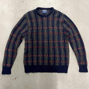 Puede incluir: Un suéter gris, azul marino y rojo estampado. El suéter tiene cuello redondo y mangas largas. El patrón consiste en rayas verticales azul marino con cuadrados rojos y rayas horizontales grises.
