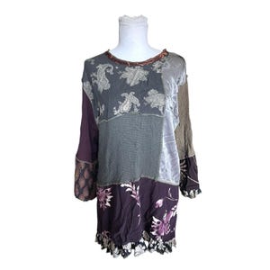 May include: A long-sleeve tunic featuring a patchwork design with various fabric textures and patterns. The garment includes sections of dark purple, grey, and brown, with paisley prints, floral motifs, and a ruffled hemline at the bottom edge.