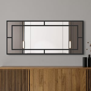 Könnte beinhalten: Rechteckiger Wandspiegel mit schwarzem geometrischen Rahmen. Der Spiegel hat eine reflektierende Oberfläche und zwei vertikale Leuchten. Der Spiegel ist an einer grauen Wand über einem Holzschrank montiert.