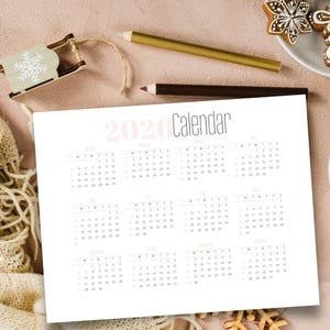 Può includere: Un calendario 2026 stampato su carta bianca, con sfondo rosa chiaro. La parola "Calendar" è scritta in un font moderno. Sono visibili anche una piccola slitta di legno e delle matite.