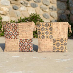 Può includere: Due borse a tracolla quadrate realizzate in sughero naturale. La borsa a sinistra presenta un motivo geometrico a rombi blu, rossi e gialli. La borsa a destra mostra un motivo floreale grigio scuro e giallo. Entrambe hanno cinturini laterali annodati.