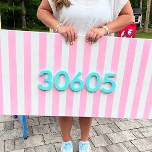 Op de afbeelding: Een rechthoekig bord met roze en witte verticale strepen en het turquoise nummer "30605". De persoon die het bord vasthoudt, draagt een zonnebril en een witte top, buiten.