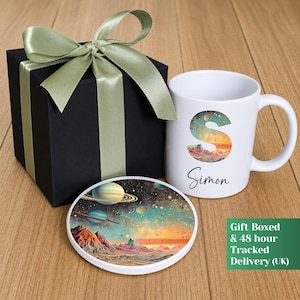 Peut inclure: Un mug en céramique blanche orné d'une grande lettre S remplie d'un motif spatial coloré, du prénom Simon en écriture cursive, et d'un dessous de verre rond assorti. L'ensemble est présenté dans une boîte cadeau noire avec un ruban vert sauge.