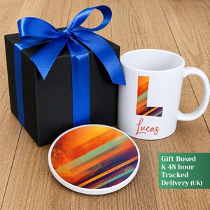May include: A white ceramic mug featuring a large letter L with a multicolored diagonal stripe pattern and the name Lucas printed below it, accompanied by a matching round coaster and a black gift box tied with a bright blue satin ribbon.