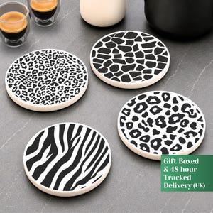 May include: A set of four round coasters featuring distinct black and white animal print patterns, including leopard spots, giraffe patches, and zebra stripes. Each coaster has a smooth, circular edge and a flat surface, arranged on a dark gray surface.