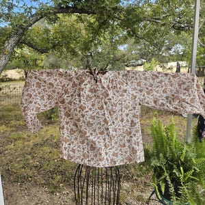 May include: A long-sleeved, off-white blouse with a brown floral pattern. The blouse is displayed on a black metal dress form. The background includes green foliage and a glimpse of a natural outdoor setting.