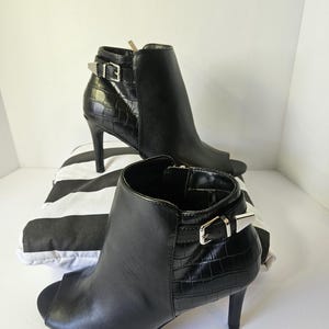 May include: A pair of black peep-toe ankle boots featuring a smooth front panel and a textured crocodile-embossed heel section. Each boot includes a side strap with a silver-toned metal buckle and a slim high heel, providing a sleek appearance.