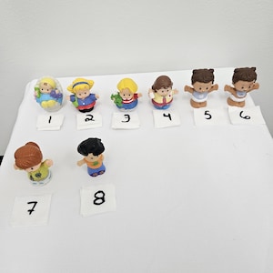 May include: A collection of eight small, colorful toy figures. The figures are of children and babies, each with unique outfits and hairstyles. The toys are numbered on small white tags. The background is a plain white surface.