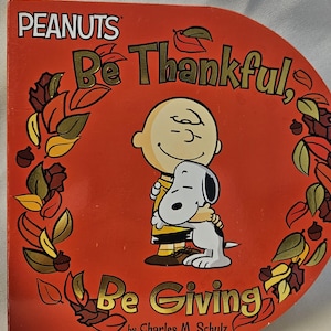 May include: A Peanuts board book titled Be Thankful, Be Giving by Charles M. Schulz. The orange cover features a central illustration of Charlie Brown hugging Snoopy, surrounded by a decorative border of autumn leaves and acorns in gold tones.