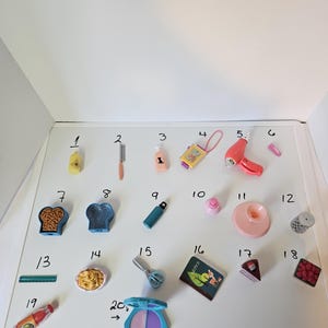 May include: A collection of miniature toys, including a yellow bottle, a knife, a pink bottle, a yellow device with a pink cord, a pink hairdryer, a pink capsule, a bread-shaped container, a blue container, a blue water bottle, a pink bottle, a pink plate, a wire basket, a green ruler, a pasta dish, scissors, a book with the word "notes", a cake slice, a tray of raspberries, a bottle of sauce, and a blue and purple makeup compact.