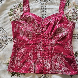 May include: A magenta-colored camisole top with white floral patterns. The top features a button-up front, wide straps, and a fitted bodice. The fabric has a subtle texture, and the floral design is prominent.