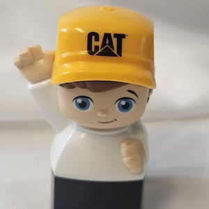 May include: A small plastic toy figure wearing a bright yellow baseball cap with the black CAT logo printed on the front. The figure has brown hair, blue eyes, a white long-sleeved shirt, and black pants, with one arm raised in a waving gesture.