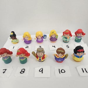 May include: A collection of eleven small plastic princess figurines. The figurines have various hair colors and wear colorful dresses. Each figurine is numbered on a small white square.