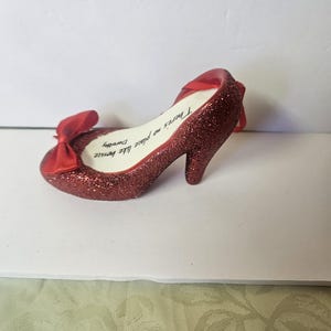 May include: A sparkly red high-heeled shoe ornament with a bow on the toe. The shoe has the text "There's no place like home Dorothy" written on the insole. A red ribbon is attached for hanging.