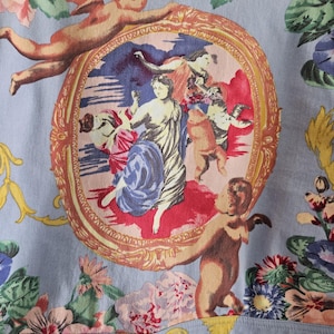 May include: Light blue fabric with a detailed print featuring cherubs, classical figures, and floral elements. The central design is a framed oval scene with figures in a classical style. The fabric is adorned with various flowers.