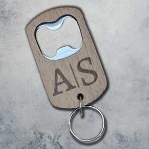 May include: A wooden bottle opener with a silver metal opener and keyring. The opener is engraved with the letters "A|S". The wood has a light brown colour.