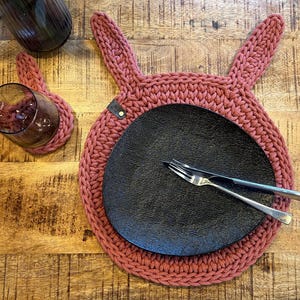 Könnte beinhalten: Ein handgefertigter, staubrosa-farbener Hasen-Platzdeckchen mit passendem Untersetzer. Ein schwarzer Teller, eine Gabel und ein Messer liegen auf dem Platzdeckchen. Das Platzdeckchen und der Untersetzer sind aus dickem Strickmaterial.