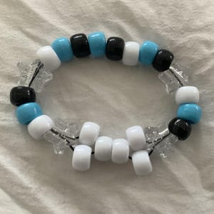 May include: A beaded bracelet featuring a pattern of white, black, and light blue beads. Clear star-shaped accents are interspersed throughout the design. The bracelet is strung on a black elastic cord.