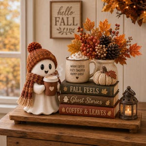 Puede incluir: Una figura de fantasma blanco con gorro y bufanda de punto color óxido, sosteniendo una taza de café marrón. Al lado, una taza moteada dice WARM COFFEE COZY SWEATER HAPPY GHOST, sobre tres libros titulados FALL FEELS, Ghost Stories y COFFEE & LEAVES.