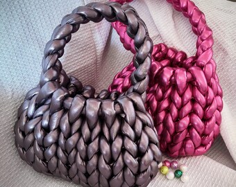 May include: Two handmade, braided handbags in metallic shades. One is silvery-grey, the other a vibrant pink. Small, colourful beads are scattered nearby. The bags have a unique, textured appearance and a braided handle.