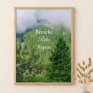 Op de afbeelding: Een ingelijste print met een weelderige groene bosscène met de woorden "Breathe. Relax. Repeat." in elegant schrift. Het kunstwerk wordt tentoongesteld boven een geweven rotan bank en een houten bijzettafel met een vaas met gedroogde bloemen.