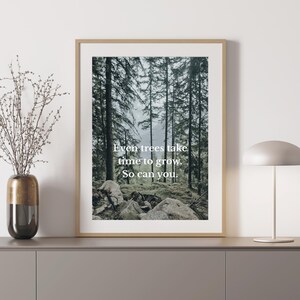 Op de afbeelding: Ingelijste kunstdruk met een boslandschap met hoge bomen en rotsen. De print heeft een licht houten frame en bevat de tekst: "Even trees take time to grow. So can you."