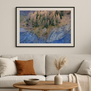 Op de afbeelding: Een ingelijste landschapsafdruk met een bos en rotsachtig terrein. Het kunstwerk toont een cluster van bomen met goudgeel blad, tegen een achtergrond van blauw getinte rotsformaties. De print wordt boven een beige bank getoond.
