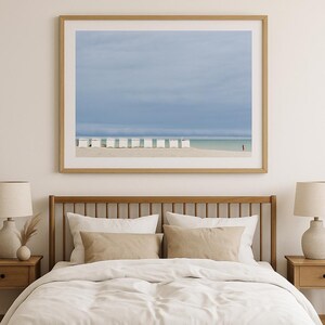Op de afbeelding: Een ingelijste prent met een strandscène onder een bewolkte hemel. Witte strandhuisjes staan langs de zandkust, met een persoon in het rood bij het water. Het kunstwerk hangt boven een bed met neutraal gekleurd beddengoed.