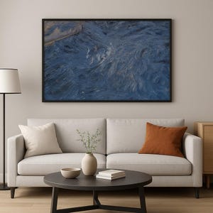 Op de afbeelding: Een ingelijst abstract kunstwerk met een zwart frame. Het kunstwerk toont een gestructureerd, wervelend patroon in tinten blauw en wit, waardoor een dynamisch visueel effect ontstaat. Het kunstwerk wordt tentoongesteld boven een lichtgekleurde bank met decoratieve kussens.