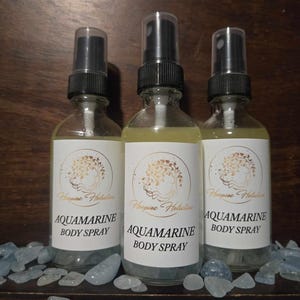May include: Three clear glass spray bottles filled with a light yellow liquid. Each bottle has a black spray nozzle and a white label with the text "AQUAMARINE BODY SPRAY". The bottles are surrounded by light blue decorative stones.