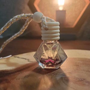 Pode incluir: Uma garrafa de vidro transparente e facetada com uma tampa de madeira e uma corda decorativa. A garrafa contém uma substância vermelha escura. A corda é branca com detalhes dourados.