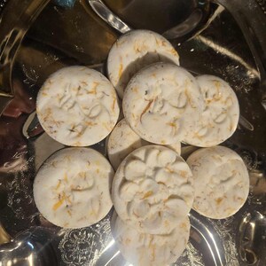 May include: A cluster of round, white bath bombs with a floral design and orange flecks, presented on a silver, ornate tray. The bath bombs have a textured surface.