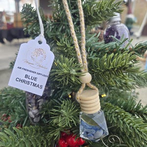 Op de afbeelding: Luchtverfrisser diffuser "Blue Christmas" hangend aan een kerstboom. De diffuser is een kleine glazen fles gevuld met blauwe stenen en een houten dop. De boom heeft groene naalden en rode bessen.