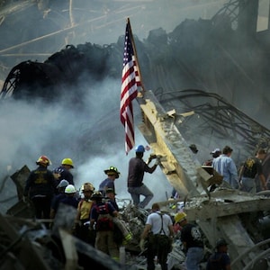 Könnte beinhalten: Eine Szene der Zerstörung mit Rauch und Trümmern. Eine amerikanische Flagge steht inmitten der Trümmer. Feuerwehrleute und Rettungskräfte sind anwesend. Die Überreste von Gebäuden sind im Hintergrund zu sehen.